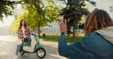 Uzun kıvırcık saçlı, kareli ceketli bir adam yeşil bir motosikletin üzerinde oturan esmer kız arkadaşının resmini çeker ve ona şehir parkında nasıl düzgün poz verileceğini gösterir..