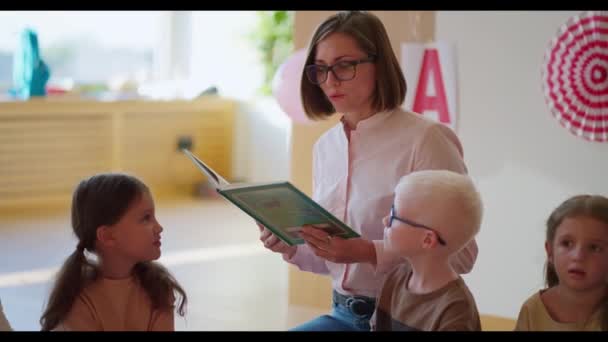 一位戴眼镜 身穿白色和粉色衬衫的金发女子为俱乐部的学生们读一本绿皮书 以帮助孩子们为上学做准备 — 图库视频影像