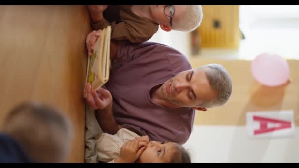 一个穿着紫色T恤衫的白发苍苍的男人为躺在地板上的孩子们朗读一本书 听他在俱乐部里为孩子们准备上学 — 图库视频影像
