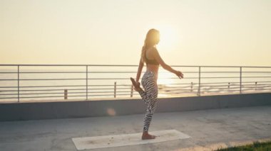 Siyah bluzlu, beyaz ve siyah pantolonlu sarışın bir su kızı yoga yapıyor ve yeşil çimlerin yanındaki kumsalda ayakta duran fiyonk çekiyor ve mavi denize ve güneşin şafağına karşı.