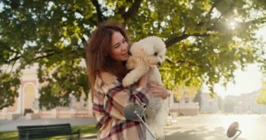 Ekose gömlekli mutlu bir esmer kız kollarında küçük beyaz bir köpek tutuyor, gülümsüyor ve mobiletinin yanındaki bir yaz parkında kameraya bakıyor..