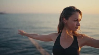 Siyah beyaz pantolonlu, siyah beyaz pantolonlu sarışın bir kız yoga yapıyor ve yaz mevsiminde gün doğumuna karşı denizin yanındaki Kızıl İskele 'de egzersiz üçgeni pozu veriyor..