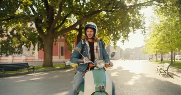 一个快乐的快递员 留着卷曲的头发 穿着牛仔夹克 骑着摩托车来到了一个城市公园 他环顾四周 在手机上看了看 想知道下一步该怎么用黄色的大头针运送他的订单 — 图库视频影像