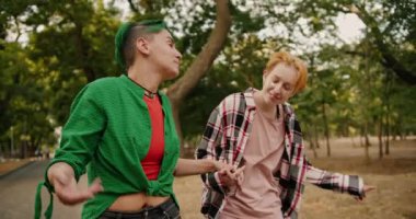 Kısa yeşil saçlı, yeşil tişörtlü lezbiyen bir kız ve ekose gömlekli sarışın kız arkadaşı iletişim kuruyor, el ele yürüyor ve parkta omuzlarını silkiyor.