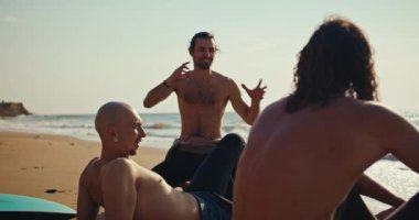 Çıplak gövdeli esmer bir adam deniz kenarındaki kumlu bir sahilde dalgıç kıyafetli bir sörfçü tarafından arkadaşlarına duygularının nasıl sörf yaptığını anlatıyor..