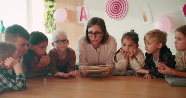 一个金发碧眼 头戴白色衬衫 戴着眼镜的女孩 在和听她讲话的孩子们坐在地板上的时候 给学龄前儿童读了一本书 — 图库视频影像
