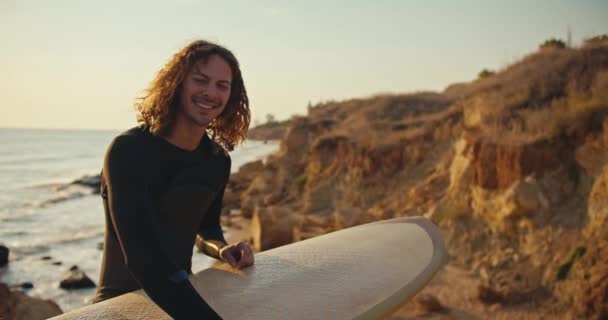 一个穿着黑色湿衣卷发的年轻人的画像 他手里拿着一块黄色的冲浪板 背景是夏日早上在日出的海滨岩石和蓝天 — 图库视频影像