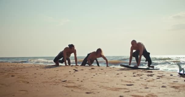 一位穿着湿衣的赤裸胸部男子向他的朋友们展示了如何在靠近大海的沙滩上正常冲浪 两个人在学冲浪 他们的朋友在帮他们 — 图库视频影像