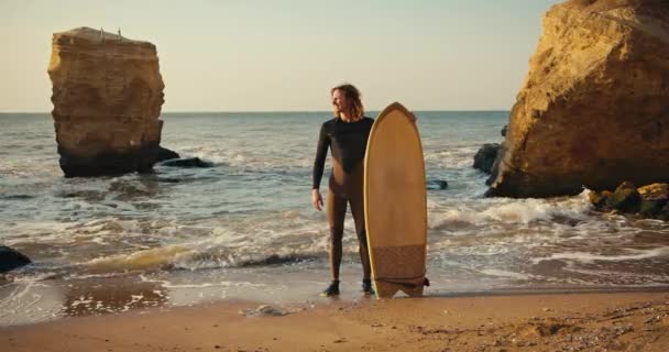 一个留着卷发的男人站在海滨 手里拿着一个浪板 浪涛汹涌 岩石汹涌 冲浪的家伙带着冲浪板沿着海岸走着 — 图库视频影像
