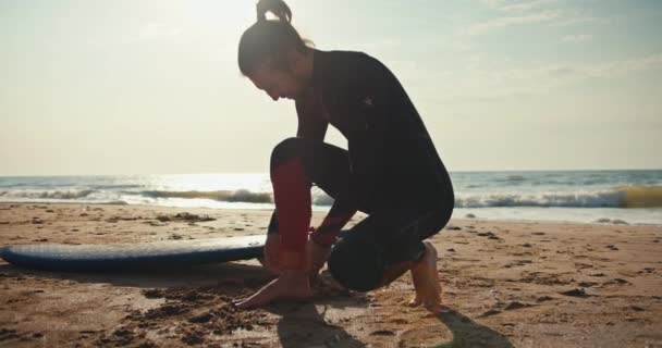 一个穿着湿衣的黑发男子在他的腿上安装了一个特殊装置 以便在海上游泳时控制他的板 男子冲浪运动员准备在海滨冲浪时游泳 — 图库视频影像