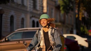 Uzun kıvırcık saçlı, kot ceketli yeşil motosikletli bir adam güzel bir yaz şehrinde yol boyunca araba sürüyor..