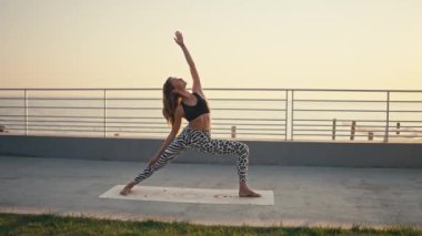 Siyah pantolonlu, beyaz pantolonlu, atletik sarışın kız yoga yapıyor ve yaz mevsiminde güneş doğarken yeşil çimlerin ve mavi denizin yanında egzersiz yapıyor..