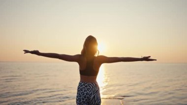 Siyah bluzlu, siyah beyaz pantolonlu genç bir atletik kız gün doğumuna karşı deniz kenarındaki bir iskelede yoga yapıyor ve meditasyon yapıyor. Fiyat meditasyonu yapan genç bir kızın arkasından bak.