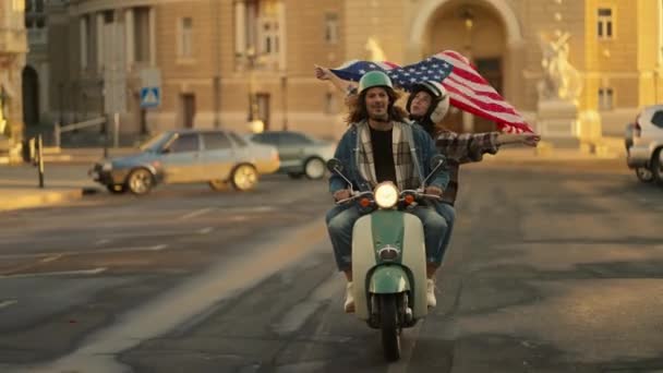 一个黑发男子穿着斜纹棉布夹克 卷曲长发 与他的女友骑在一起 女友手里拿着一面美国大旗 在他们乘坐夏季城市的途中 飘扬在风中 — 图库视频影像