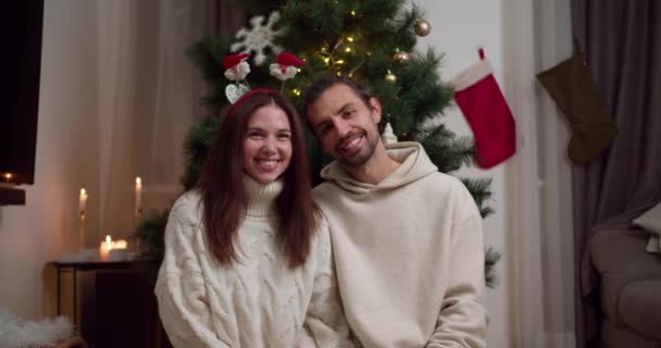 一对快乐的夫妇的画像 一个穿着白色毛衣的黑发男人和一个穿着白色毛衣的黑发女孩在温暖的冬宫里摆姿势看着他们圣诞树旁的相机 — 图库视频影像