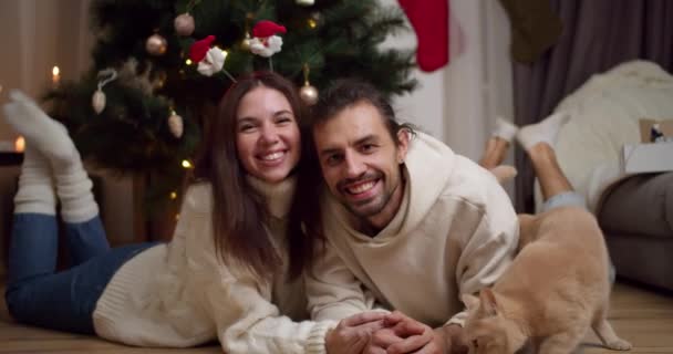 一对快乐的夫妇 一个穿白衫的男人和一个黑发女孩的画像躺在圣诞树旁他们那只乳白色的猫旁边的地板上 — 图库视频影像