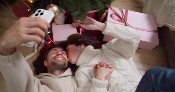 ブルネットの男と白いセーターの彼のガールフレンドは床に横たわり 冬の居心地の良い部屋で新年の木とピンクの贈り物の近くにセルフィーを取ります — ストック動画