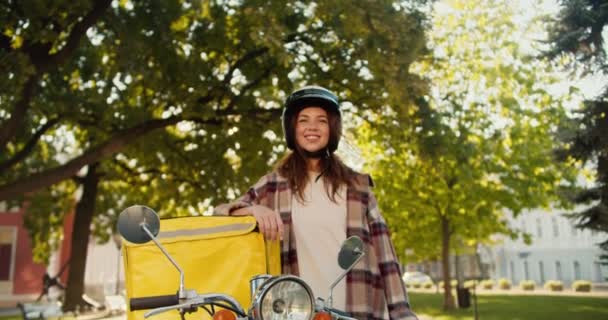 ハッピーブルネットの宅配女の子の肖像画 チェッカーのシャツに白いオートバイのヘルメットを着て 夏のストリートでの食糧配達のための黄色いバッグ — ストック動画