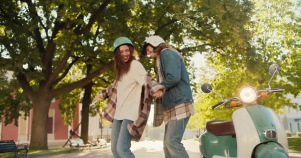 市内の晴れた夏の公園で彼女の緑の近くのデニムジャケットで彼女のボーイフレンドと踊るデニムパンツのハッピーなシャツとオートバイのヘルメットでハッピーガール — ストック動画
