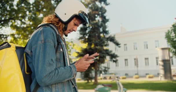 一个穿着斜纹棉布衬衫卷发的快递员穿过一个夏季的公园 环顾四周 看了看他的手机 然后骑上他的摩托车 提着一个黄色的大包 离开去送他的东西 — 图库视频影像