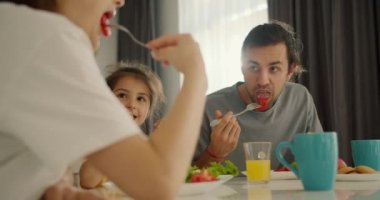 Yakın plan çekim: mutlu esmer adam kahvaltıda küçük kızıyla birlikte salata yiyor. Karısından aldığı sarı elbiseyle beyaz tişörtlü modern bir dairede beyaz bir masada.