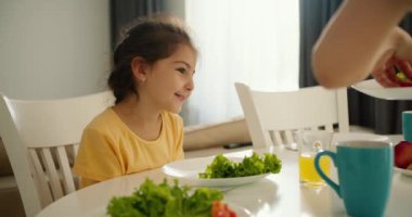 Sarı elbiseli küçük esmer bir kız mutfaktaki sandalyeye oturur ve annesi sabah kahvaltısı için kızın tabağına yeşil otlar ve sebzeler koyar..