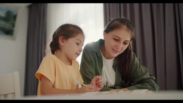一个穿着黄色衣服的褐发小女孩和她的妈妈一起做作业 一个穿着绿色夹克的褐发女人 坐在舒适的现代公寓的桌子边 一边在笔记本上写字 — 图库视频影像