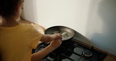 Sarı elbiseli esmer bir kızın, annesiyle birlikte tavuğa yumurta kırarken ve modern bir mutfakta omlet pişirirken görüntüsü..