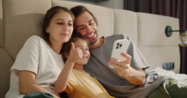 Gri tişörtlü esmer bir adam, karısı ve sarı elbiseli esmer kızıyla birlikte modern bir dairede açık kahverengi bir yatakta beyaz bir telefonla selfie çekiyor..