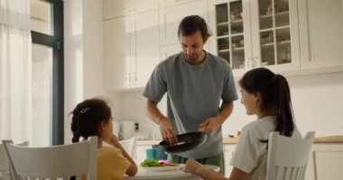 Gri tişörtlü esmer bir adam, sabah aile masasındaki mutfakta kahvaltı ederken karısı ve küçük kızı için kızartma tavasından sıcak tost serer..