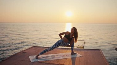 Siyah beyaz pantolonlu, siyah pantolonlu, poz veren genç sarışın kız denizin karşı tarafında kırmızı ahşap bir iskelede yoga yaparken ve yazın gündoğumunda egzersiz yapıyor..