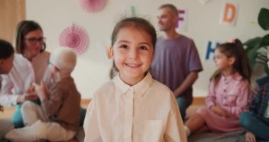 Portre: beyaz gömlekli kahverengi gözlü mutlu küçük kız kameraya bakıyor ve anaokulu kulübündeki ilk dersine gülümsüyor. Küçük kız okuldaki ilk dersinde oturuyor.