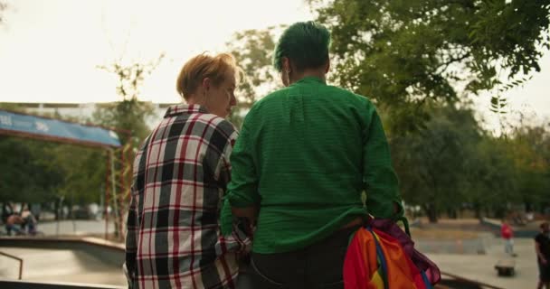 在他们的约会中 一个绿头发 绿衬衫短发的女孩和一个格子衬衫短发的金发女孩坐在一个溜冰场的篱笆上 她们正在和别人聊天 — 图库视频影像