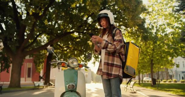 一位身穿格子衬衫 头戴白色摩托车头盔 头戴黄包的黑发女信使站在摩托旁边 她看着手机屏幕 想知道在哪里下订单 — 图库视频影像
