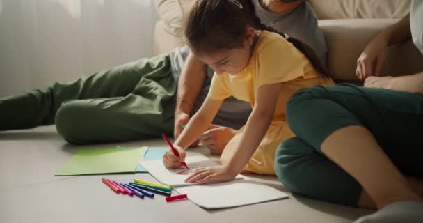 在现代公寓里 一个穿着黄色衣服的金发小女孩坐在父母身边的地板上 用彩色毛笔在纸上画画的特写镜头 — 图库视频影像