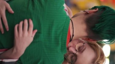 Dikey video: Kısa saçlı kibar sarışın bir kız eğlence parkındaki randevusu sırasında kısa yeşil saçlı kız arkadaşına sarılıyor..