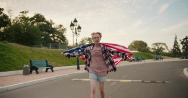 Kısa saçlı, gözlüklü, pembe gömlekli sarışın bir kız yazın parktaki yolda omuzlarının arkasında Amerika Birleşik Devletleri bayrağıyla koşuyor..