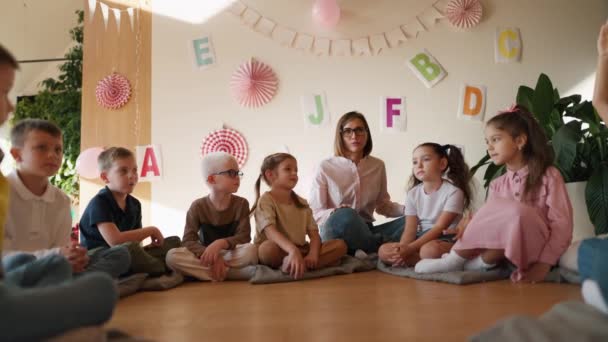 メガネのボブヘアスタイルと白いシャツを持つ幸せなブロンドの少女は 学校のために子供を準備し 通信するためにクラブの部屋の学生と特別な枕の上に床に座っています — ストック動画