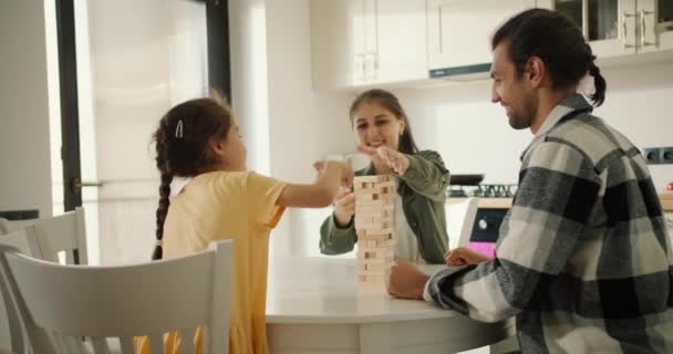 一个穿着黄色连衣裙的黑发小女孩和她的父母在一个白色的现代化厨房的桌子上玩棋盘游戏Jenga的后视镜 — 图库视频影像