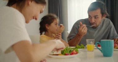 Kirli sakallı esmer bir adam beyaz tişörtlü karısıyla kahvaltı eder ve küçük kızı da sabah modern bir dairede aile masasında sarı bir elbise giyer..