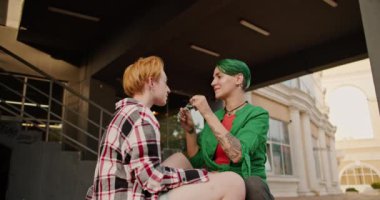 Ekose gömlekli kısa saçlı iki lezbiyen kız büyük bir binanın yanında oturuyor. Yeşil tişörtlü, kısa saçlı lezbiyen kız gözlüklerini çıkarıp sarışın kızına sarılıyor.