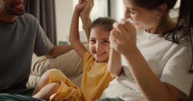 Yakın planda beyaz tişörtlü esmer bir anne, açık kahverengi bir kanepede babasıyla modern bir stüdyo odasında otururken sarı elbiseli kızıyla oynuyordu..