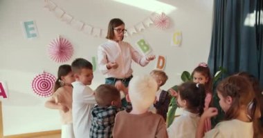 Kısa saçlı, beyaz gömlekli ve kot pantolonlu sarışın bir öğretmen kız anaokulu çocuklarına kollarını nasıl gereceklerini ve kısa ve sıcak bir şekilde nasıl hareket edeceklerini anlatan bir ders veriyor.