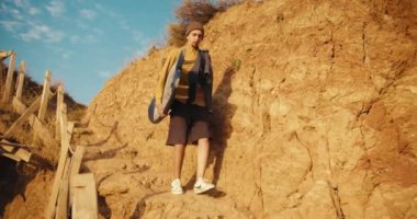 Turuncu tişörtlü bir adam kayalık deniz kıyısına gider ve güneşli bir yaz sabahında elinde sörf tahtası taşır. Şapkalı adam yazın sörf yapmak için sörf tahtasıyla denize gider..