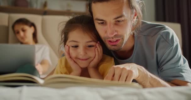 一位穿着灰色T恤的黑发男子正和他那穿着黄色T恤的感兴趣的小黑发女儿一起看书 而她的母亲则躺在沙发上 躺在笔记本电脑上休息 — 图库视频影像