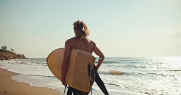 看到一个穿着湿衣赤身裸体的快乐的卷曲冲浪手在沙滩上奔跑 手里拿着冲浪的照片 在夏日的海面上滑行 — 图库视频影像