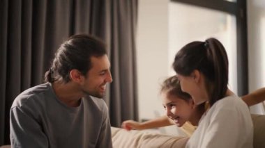 Gri tişörtlü esmer bir adam ve beyaz tişörtlü esmer bir kız, küçük kızlarına sarılıyorlar, sarı elbiseli esmer bir kız kanepeye atlıyor ve ona sarılıyor.