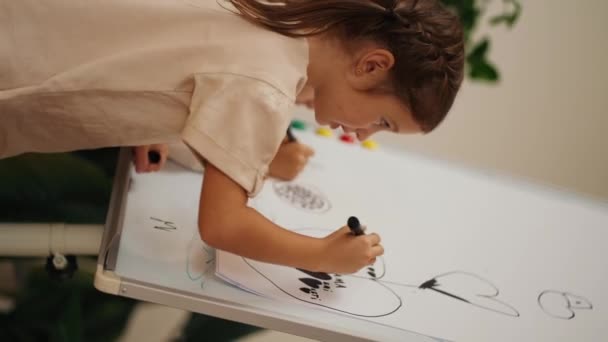 一个穿着白色T恤衫的女孩的垂直录像 上面有一张大纸片上的黑色记号 装在俱乐部房间的磁板上 为学龄前儿童入学做准备 — 图库视频影像