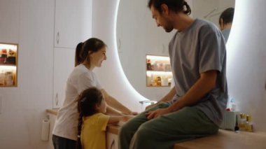 Gri tişörtlü esmer bir adam banyodaki masanın üzerine oturur ve modern bir banyonun aynasının yanındaki modern lavaboda karısıyla kızının ellerini yıkamasını seyreder..