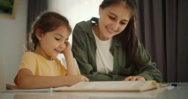 Sarı tişörtlü esmer bir kız ödevini yapar ve defterine yeşil ceketli esmer bir kız ve beyaz tişörtlü bir kızla birlikte yazar.